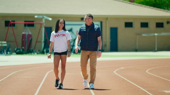Two individuals walk alongside each other on a red, white-lined track. The person on the left is wearing a white shirt black shorts, and black shoes, while the person on the right is wearing a navy vest and shirt, khaki pants, and white shoes. A brown building sits in the background.