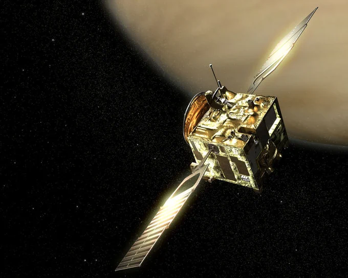 The picture shows an artist's impression of Venus Express orbiting the planet Venus. The spacecraft is the shape of a cube and is a golden colour. Two solar panels extend outwards on two sides, the light of the sun is reflected on the panels. Venus can partially be seen in the background, in the top right-hand corner of the image. The surface of the planet is a beige colour.