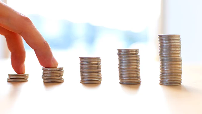 Five stacks of coins in a row on a table, gradually taller from left to right. A person's two fingers are on the two leftmost stacks.