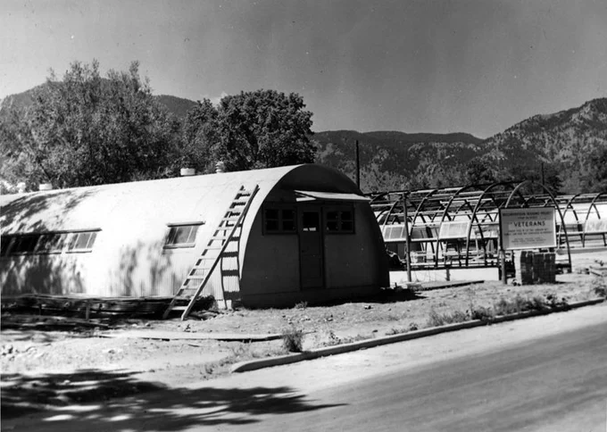 Black and white photo of a Quonset hut with two additional huts under construction in the background. Mountains and trees beyond.