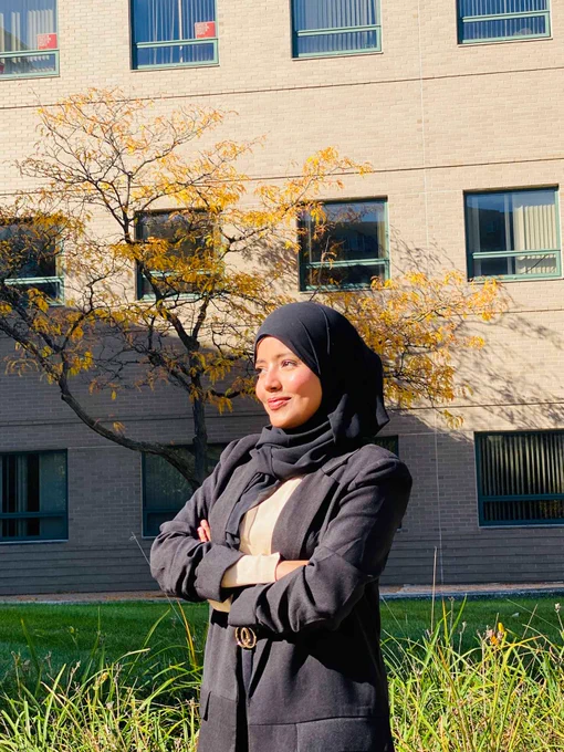 Person in a black outfit and hijab standing confidently with arms crossed in front of a school building with a tree displaying autumn foliage.