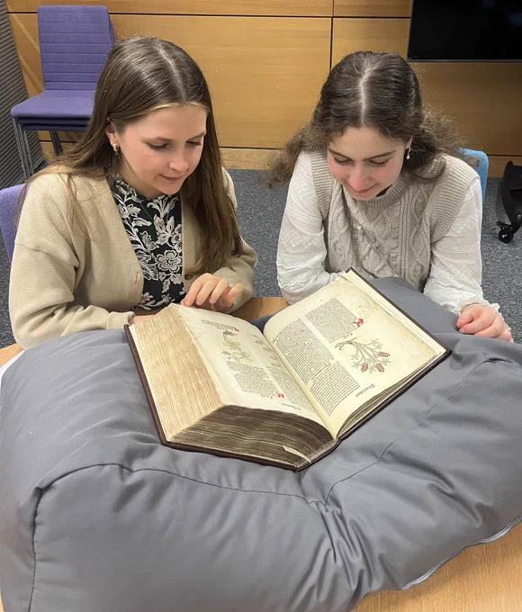 Two young white women looking at the Ortus Sanitatis, a 15th century natural history encyclopaedia, on a grey book cushion.