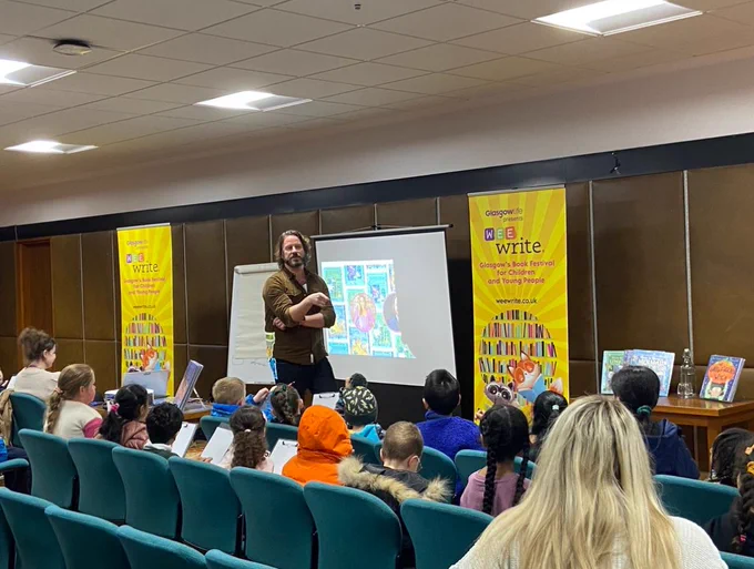 Illustrator Steve Lenton standing at front of lecture room talking to group of children. Backdrop is large screen and yellow display panels with Wee Write logo. 