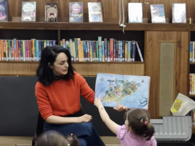 Author Helen Kellock reading book, showing pages with picture of an island. A young girl is pointing to the image. The background is library shelves