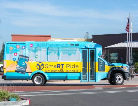 Photo credit: SacRT. Photo of Sacramento Regional Transit's SmaRT Ride on-demand transit vehicle, colored blue with a yellow stripe. The vehicle is in front of a building complex. 