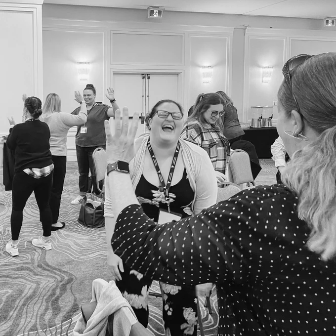 An attendee in glasses smiles wide at a colleague. Educators are in the back doing exercises related to a topic of discussion.