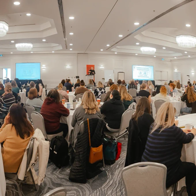 A large group of attendees listens to a speaker on stage at the conference.

