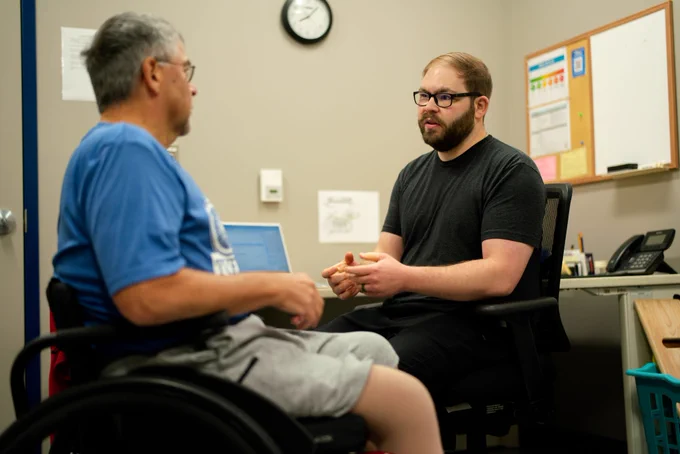 A man in a wheelchair talking with another man sitting at a desk. The man sitting at the desk is a physical therapist. 