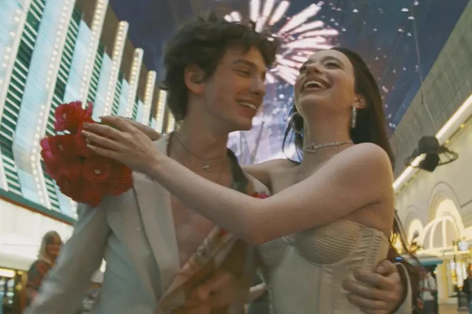A man in a white suit wraps one arm around the waist of a woman in a white sleeveless dress. The woman’s arms are wrapped around the man’s shoulders, with a bouquet of roses in one hand. Both of them are smiling. Courtesy of NEON.
