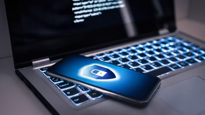 A photo of a smartphone with an image of a lock and shield on the screen resting on the keyboard of a laptop. 