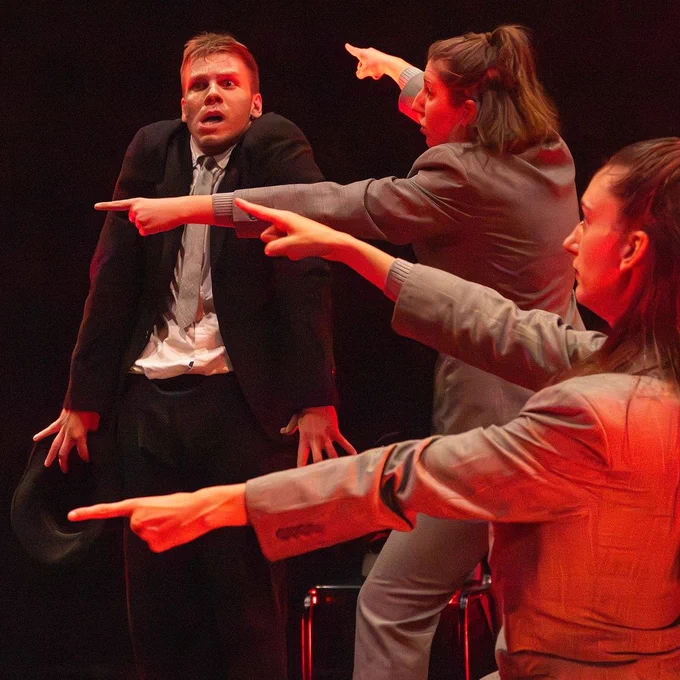 Three actors on stage in business attire performing a dramatic scene, with expressive gestures pointing at the central figure who looks surprised.