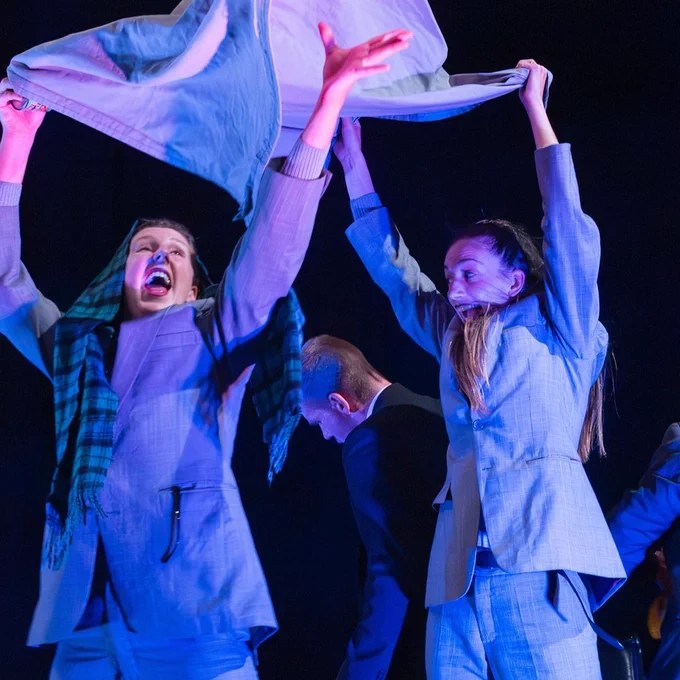 Two performers in business suits joyfully waving fabrics above their heads on a stage with dramatic lighting.
