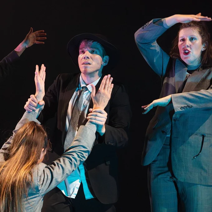 A dynamic theater scene featuring four performers; one in a suit and hat is at the center, surrounded by others gesturing dramatically towards them under a spotlight.