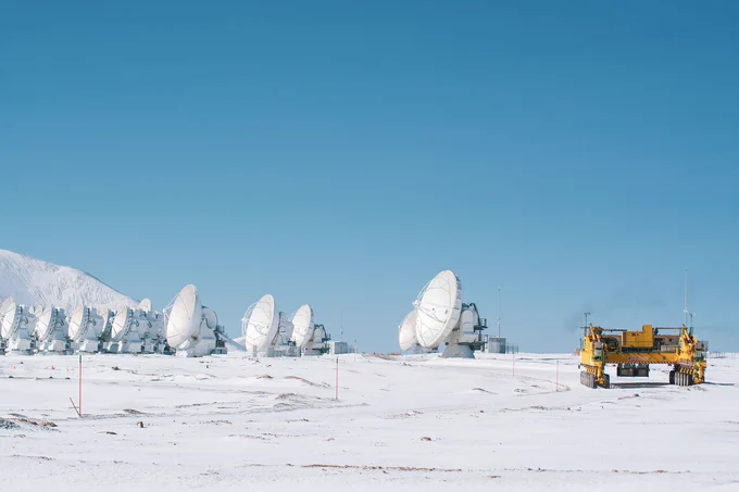 Eng beieinanderstehende weiße Antennenschüsseln in einer schneebedeckten Landschaft unter blauem Himmel, im Vordergrund ein gelbes Transportfahrzeug mit vielen Rädern