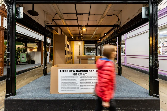 A blurry red coated figure in front of Leeds Low Carbon Pop up, an installation in Kirkgate Market built from cardboard
