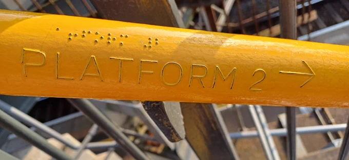 Bright yellow handrail with raised letters reading platform 2 with arrow pointing the way below braille 