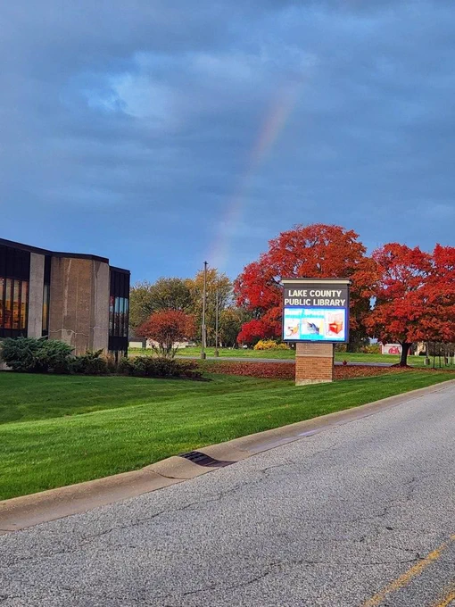 A rainbow beaming down from the sky between the Merrillville Branch sign and the building. The trees behind the sign are brilliantly red