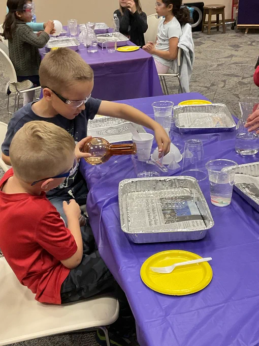 wo kids wearing safety goggles slowly pour a liquid out of an orange beaker into a measuring cup