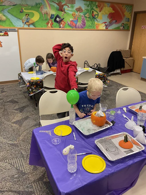 A child looks astonished at a self-inflating balloon while another peers closely at a pumpkin in a tray of bubbling suds