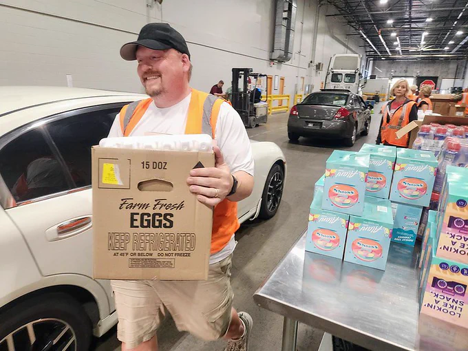 A person wearing a cap is smiling while carrying a large box labeled "Farm Fresh Eggs" in a warehouse with vehicles and other packaged foods around.