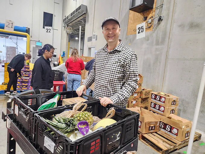 Individual volunteering at a food bank, organizing crates of fresh produce, wearing a plaid shirt and a cap, with other volunteers working in the background.