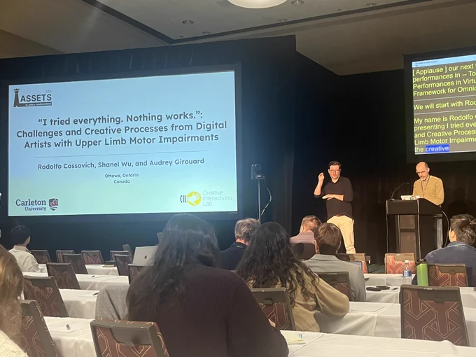 The photo is taken at a presentation room with two digital screens. Presenter is facing the audience and main screen shows the first slide with a title heading “I tried everything. Nothing works: Challenges and Creative Processes from Digital Artists with Upper Limb Motor Impairments”.