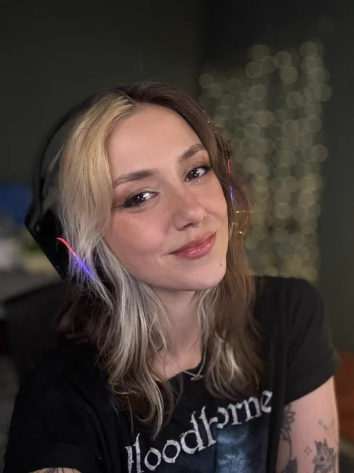 Ellie is smiling in a selfie in her stream room. She is wearing her shoulder length brown and blonde hair loose and wavy. She is wearing a black Bloodborne t shirt, black over ear headset, and has illustrative linework tattoos on her arms