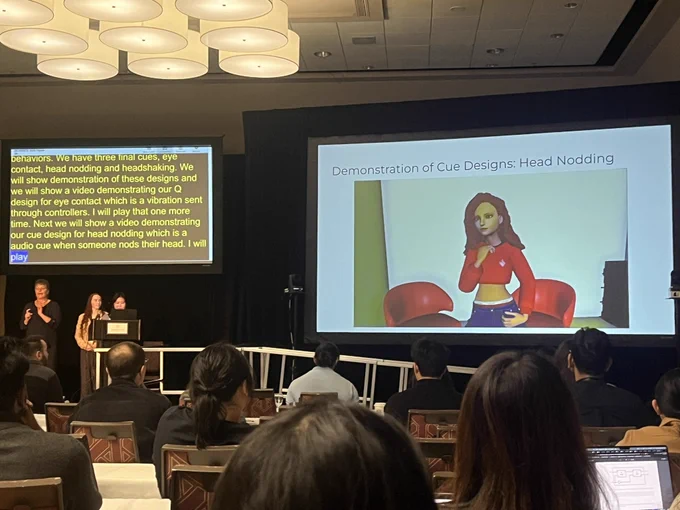 Photo is taken at a conference room with two digital screens. Two presenters and a sign-language interpreter are on the stage. A virtual reality avatar is head nodding on the first screen, and subtitles are on the second screen.