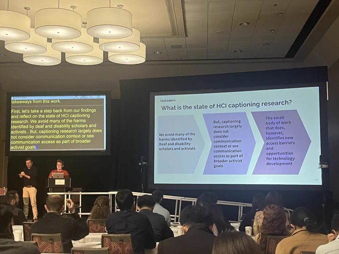 Photo is taken at a conference room with two digital screens. A presenter and a sign language interpreter are on the stage. A slide with a heading “What is the state of HCI captioning research” is on the first screen and subtitles are on the second screen.