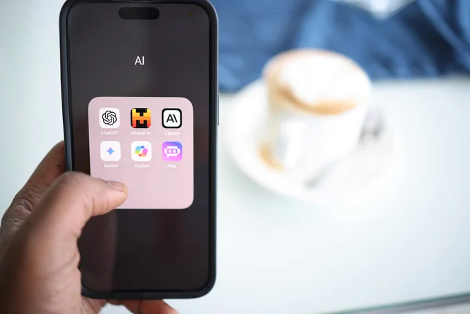 A person holding an iPhone in the left hand, showing a folder with the name "AI" that includes apps like chatGPT, Copilot, Claude and other generative AI services. The background is a latte on a table. Photo by Solen Feyissa on Unsplash