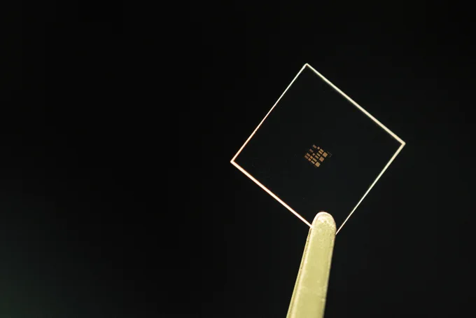 A pair of tweezers holds up a small glass square against a black background. In the center of the glass, there is a grid-like pattern of tiny sensors. Photo credit Andrew Brodhead.