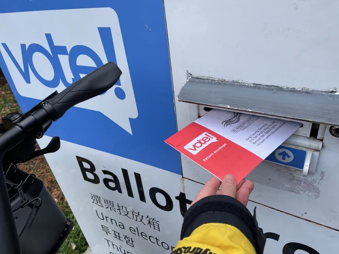 The image is a photograph of an outstretched hand inserting a ballot into a blue and white ballot box that says "vote!". The ballot is in a secure red envelope. At the left of the photo are the handlebars of a bicycle.