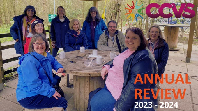 We see a group of people sat outside in a country park at a wooden table. Six people are sat down and three stood at the back. Everyone is smiling and looking at the camera. They are all wearing coats and have mugs on the table in front of them. We see the CCVS logo in the corner of the image and the words: Annual Review 2023-2024.