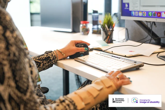 Close-up on a professional's hands as they work sitting at a desk. They are wearing a wrist support on the right side and are using an adaptive keyboard and ergonomic mouse. The Business Disability Forum 'Changing the image of disability' logo is on the lower right corner of the image.