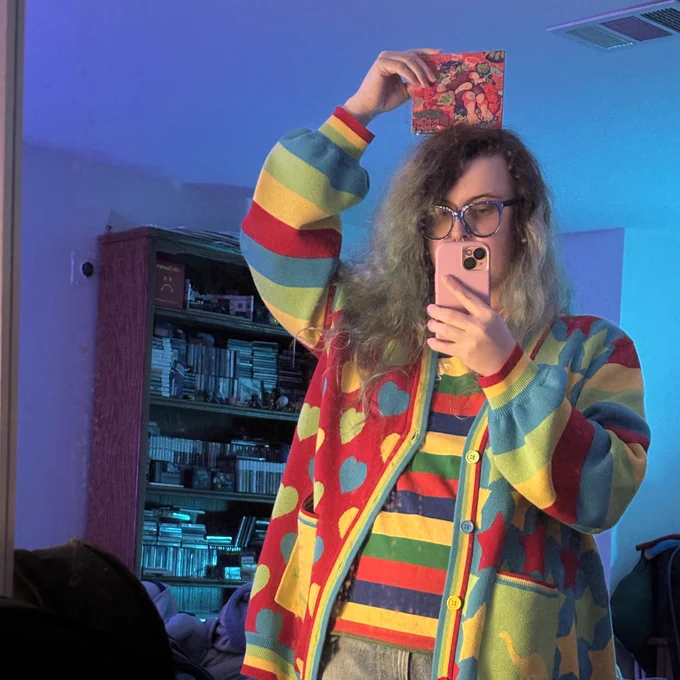 sophia (real) holds a cd on their head while taking a mirror selfie. they are wearing a colorful cardigan and shirt underneath it