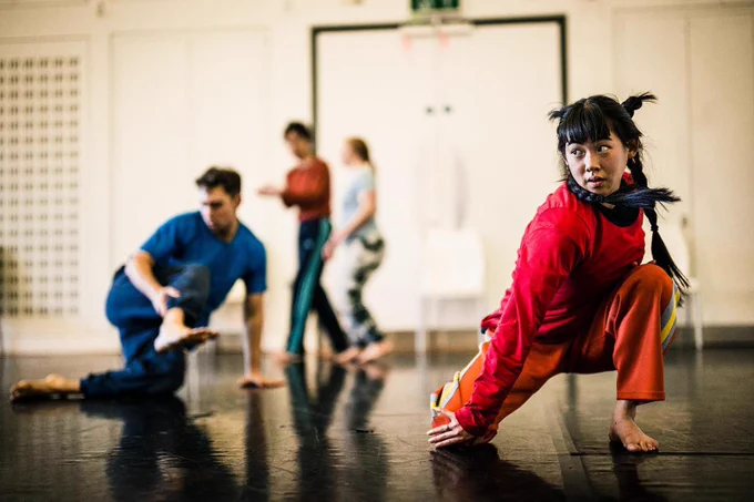 Emily from Stopgap leading a workshop. She's dressed in a red tracksuit and is crouched down with one leg stretched out to the side]