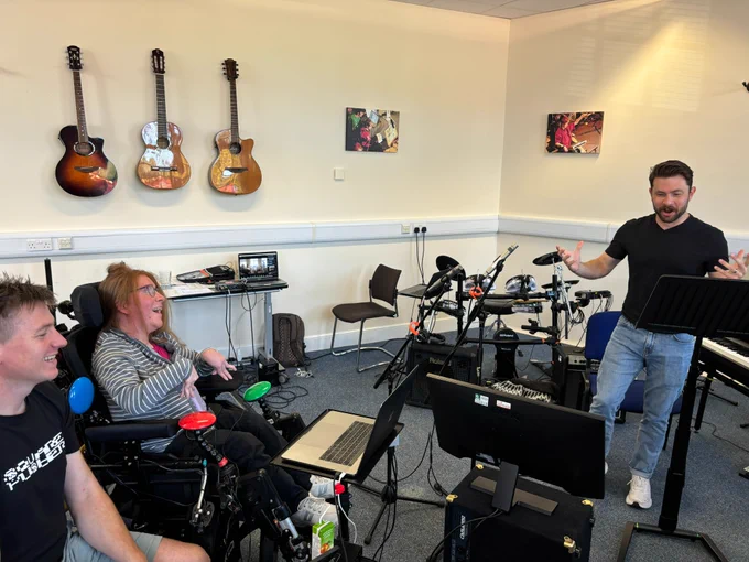 Rhona and Ali (on the left) look towards Colin, giggling. The room is full of assorted music and technological devices