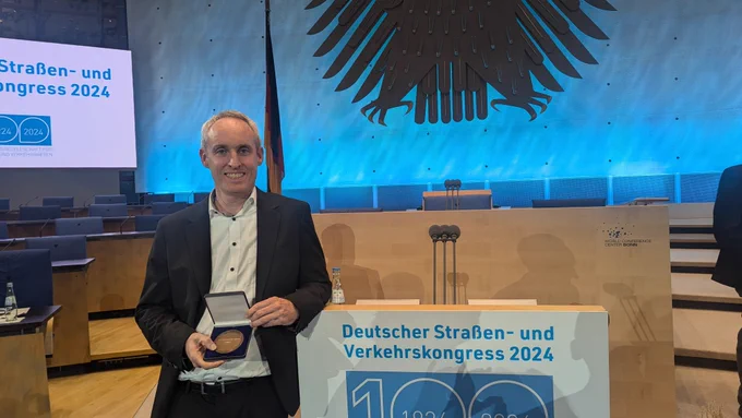 Mann im Anzug steht auf einer Bühne und hält Schatulle mit einer Münze in den Händen