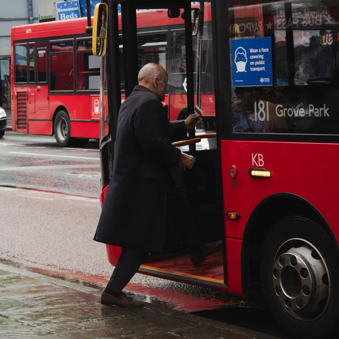 Man in a black coat holding a bus ticket, standing beside a red double-decker bus on a sunny day.