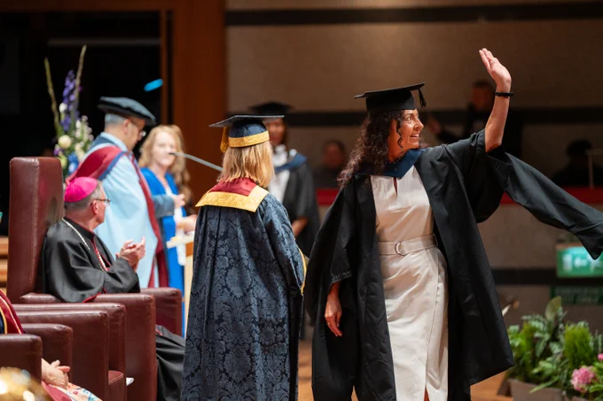 Student dressed in Graduation attire, standing on stage and waving