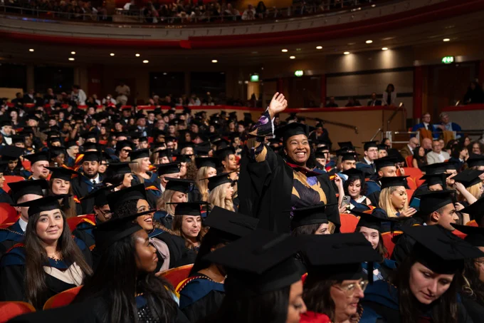 Student dressed in Graduation attire within Symphony Hall, standing in the crowd and waving
