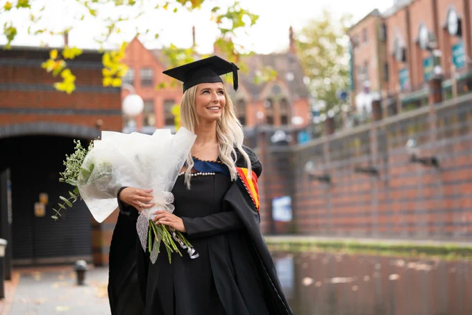 Student dressed in Graduation attire, standing outside with flowers in hand
