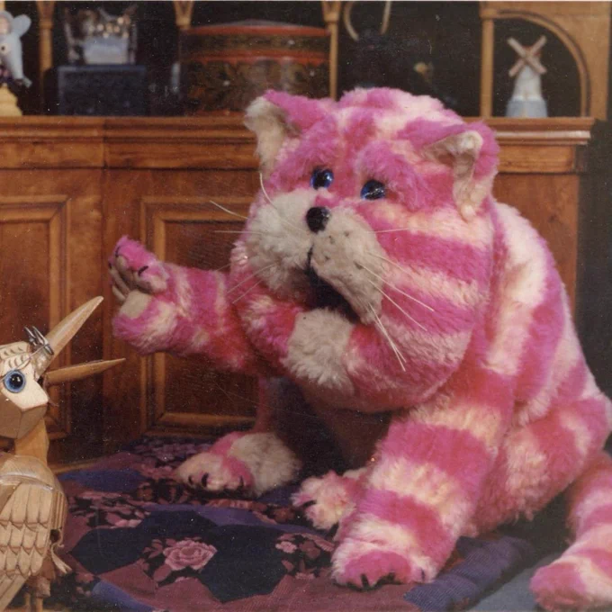 Bagpuss, a pink striped soft cat puppet with blue eyes. He raises a paw as he talks to Professor Yaffle, a wooden woodpecker puppet with glasses,