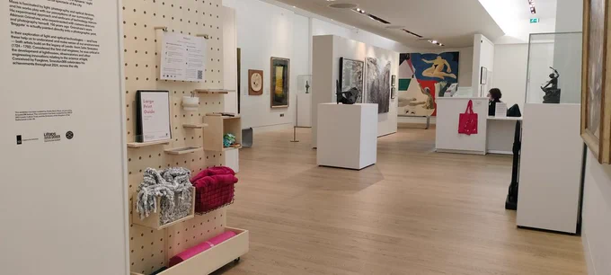 The interior of The Stanley & Audrey Burton Gallery with various paintings on white walls. In the foreground is a wooden pegboard shelving unit filled with access resources.