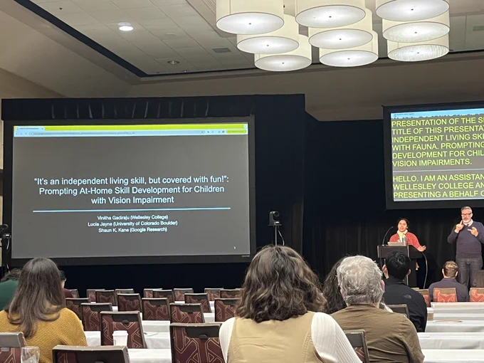 Photo is taken at a conference room with two digital screens. A presenter and a sign language interpreter are on the stage facing the audience. A title slide with a heading “It's an independent living skill, but covered with fun!": Prompting At-Home Skill Development for Children with Vision Impairment” is on the first screen and subtitles are on the second screen.