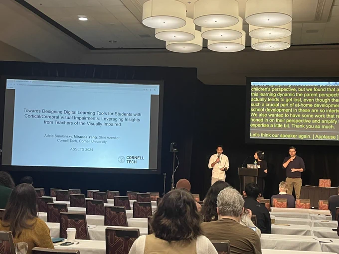 Photo is taken at a conference room with two digital screens. A facilitator, a presenter and a sign language interpreter are on the stage facing the audience. A title slide with a heading “Towards Designing Digital Learning Tools for Students with Cortical/Cerebral Visual Impairments: Leveraging Insights from Teachers of the Visually Impaired” is on the first screen and subtitles are on the second screen.