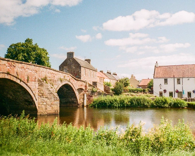 Colour photograph of Haddington's Nungate bridge, river Tyne, and old buildings on a sunny summer day
