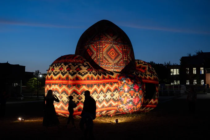 A photograph of a large, beaded sculpture at dusk. The sculpture is covered in a pattern evoking Middle Eastern rugs. The sculpture is lit from below. 