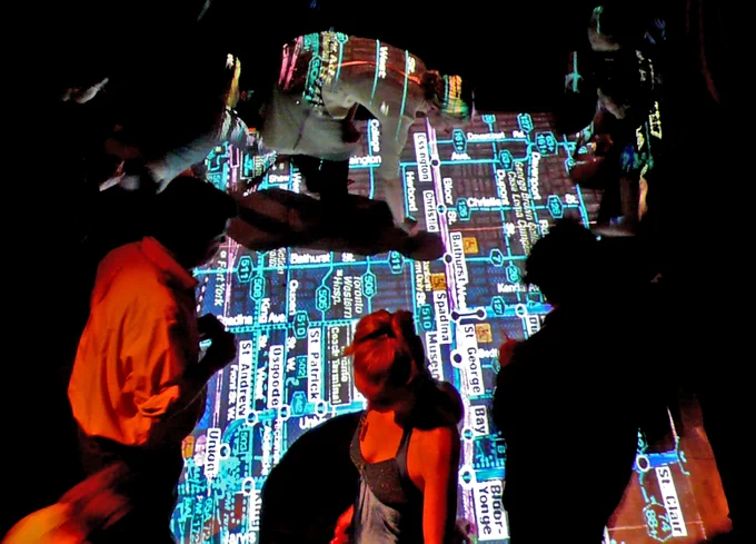 A photograph of a small group of people walking on and engaging with a projection of Toronto’s transit map. 