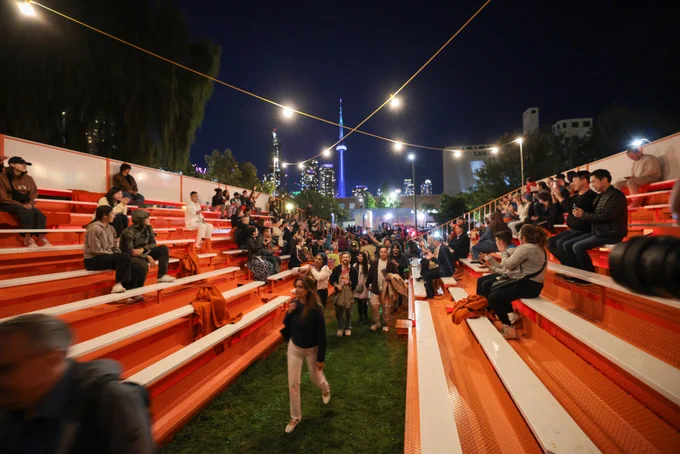 A photograph of a group of people sitting on sets of bleachers facing each other. People walk along the walkway in the centre of the two bleachers. 